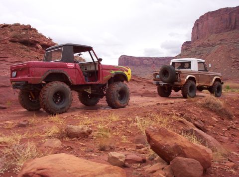 1974 Ford Bronco - Two Ford Broncos, one red and one brown, navigate a rocky desert terrain under a cloudy sky.