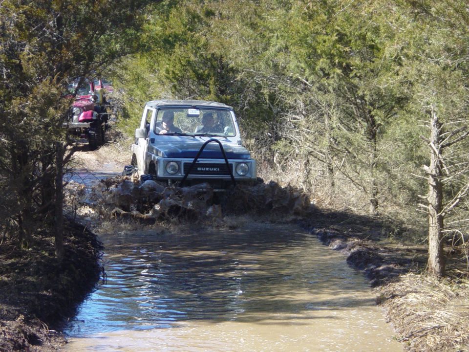 White and blue Suzuki Samurai fording muddy water on a tree-lined trail, red vehicle following behind