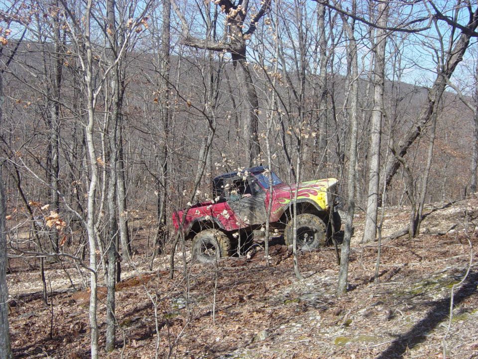 Red early Ford Bronco with yellow flames and big mud tires parked on a leafy wooded trail