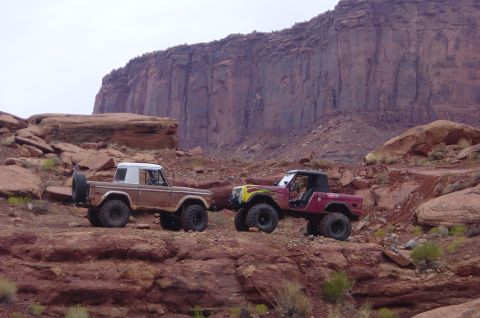 1974 Ford Bronco - Two 1974 Ford Broncos navigate a rocky desert trail with a massive cliff in the background.