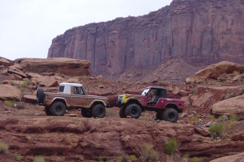 Two 1974 Ford Broncos parked on rocky terrain with a large cliff face in the background