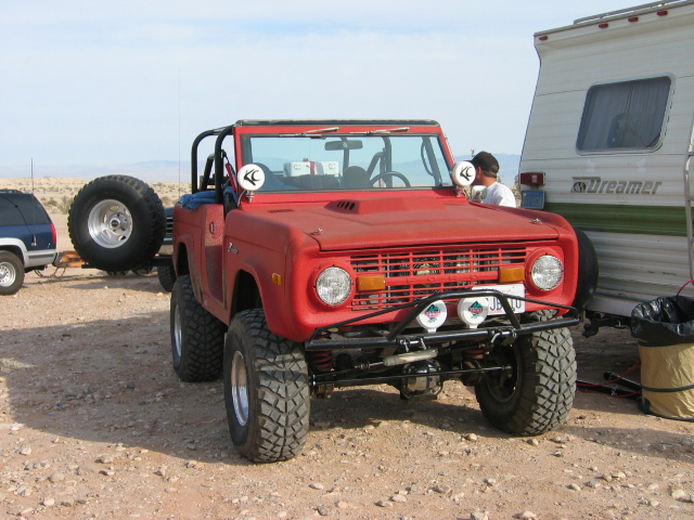 Red doorless lifted Ford Bronco on big tires parked in desert next to a camper trailer