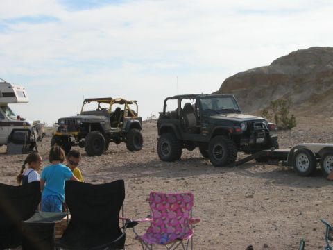 1985 Ford Bronco - Off-road vehicles and an RV are parked in a desert area with people and camping chairs nearby.