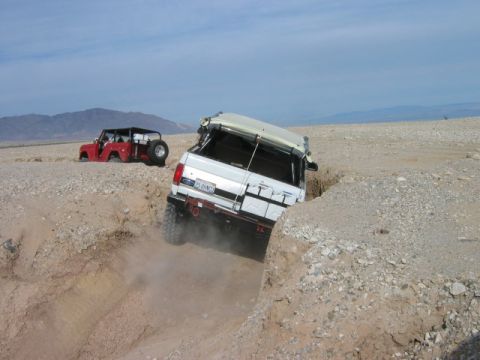 1985 Ford Bronco - Two off-road vehicles tackle a challenging desert terrain, with one vehicle stuck in a deep ditch.