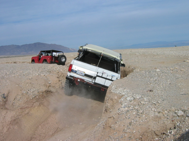 White Ford Bronco climbing into a dirt gully with red Jeep in desert background