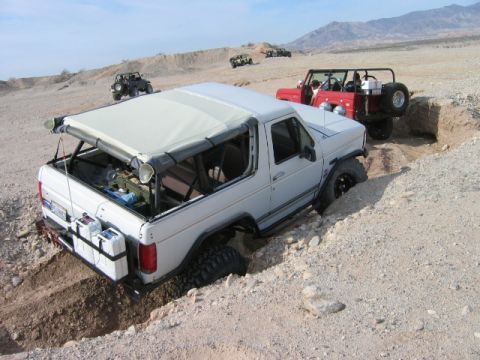 1985 Ford Bronco - Several off-road vehicles navigate a challenging desert landscape with sandy terrain and deep ditches.