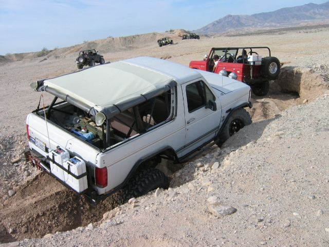 White Ford Bronco rear 3/4 view on desert trail with red Jeep ahead
