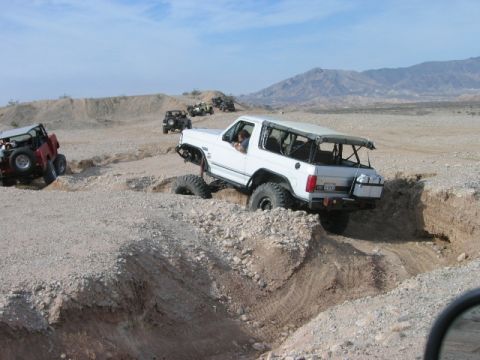 1985 Ford Bronco - Several off-road vehicles, including a white 1985 Ford Bronco, traverse a challenging desert track.