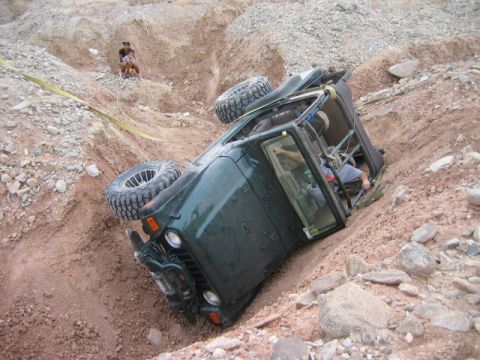 1985 Ford Bronco - An off-road vehicle has overturned in a steep dirt embankment, with a spare tire visible on its roof.