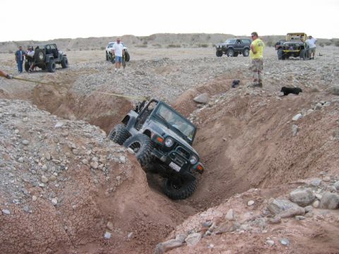 1985 Ford Bronco - Several off-road vehicles are present in a rocky, desert terrain, with one Jeep attempting to navigate a steep trench.