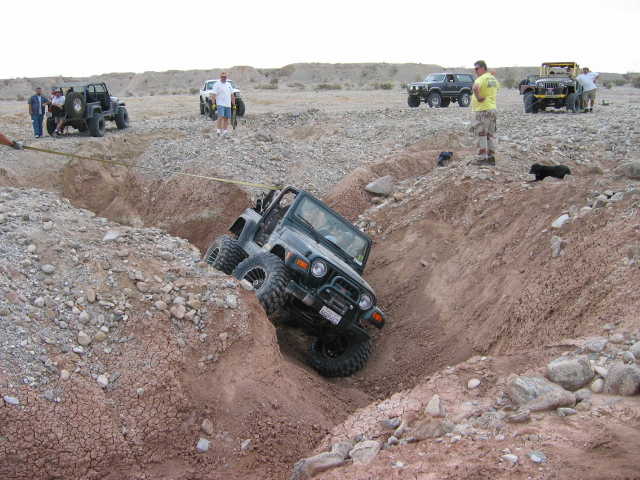 Dark Jeep on off-road tires crawling into a deep dirt gully with spectators and other Jeeps above