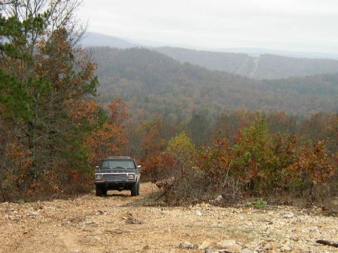 1995 Ford Bronco - A black Ford Bronco is parked on a dirt road surrounded by trees with autumn foliage, with rolling hills in the distance.