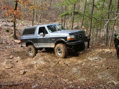 1995 Ford Bronco - A two-tone 1995 Ford Bronco navigates a challenging muddy path through a wooded area.
