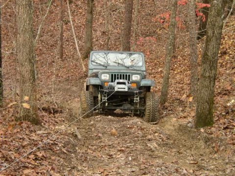 1995 Ford Bronco - Green Jeep Wrangler navigating a muddy trail in a wooded area, with a tow rope attached.