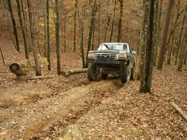 1995 Ford Bronco driving on a dirt path in a forest with fallen leaves