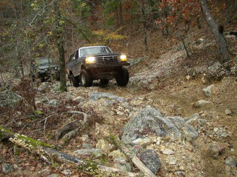 1995 Ford Bronco - Two SUVs navigate a challenging, rocky path through a wooded area during autumn.