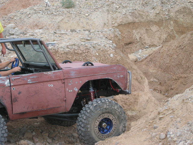 Faded red early Ford Bronco crawling a dirt pit with coilover and blue beadlock wheels