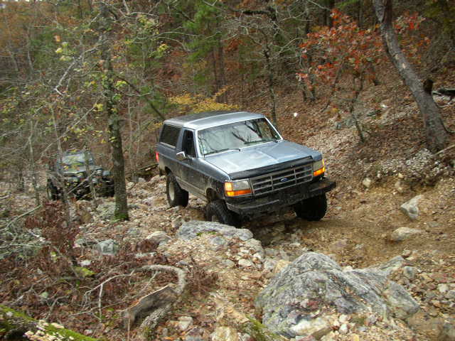1995 Ford Bronco driving on a rocky, wooded trail. Another off-road vehicle is visible in the background