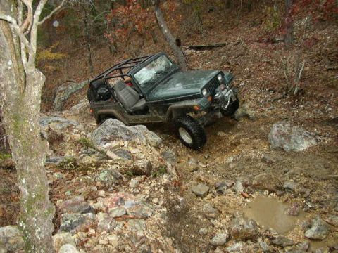 1995 Ford Bronco - A green Jeep Wrangler tackles a challenging off-road course with rocky obstacles and muddy puddles.