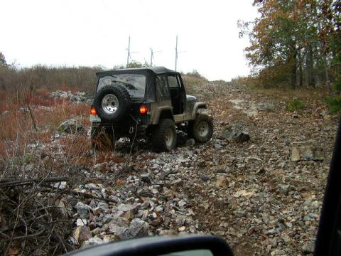 1995 Ford Bronco - Jeep Wrangler navigates a challenging rocky trail with trees and power lines in the background.