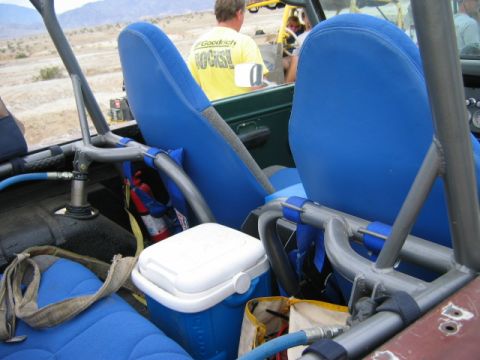 1985 Ford Bronco - Interior of an off-road vehicle with blue seats, roll cage, cooler, and fire extinguisher. A person in a yellow shirt is visible.