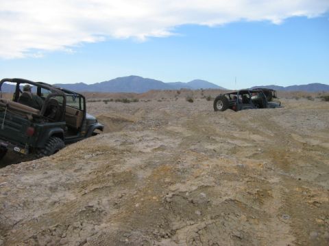 1985 Ford Bronco - Two off-road vehicles traverse a desert landscape under a partly cloudy sky.