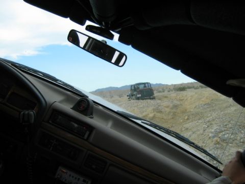 1985 Ford Bronco - View from inside a vehicle looking out at another off-road vehicle traversing a dry, rocky landscape under a clear blue sky.