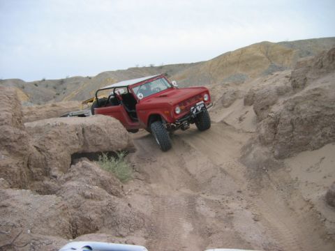 1985 Ford Bronco - Red Ford Bronco navigating a steep, sandy, off-road trail with rocky terrain.