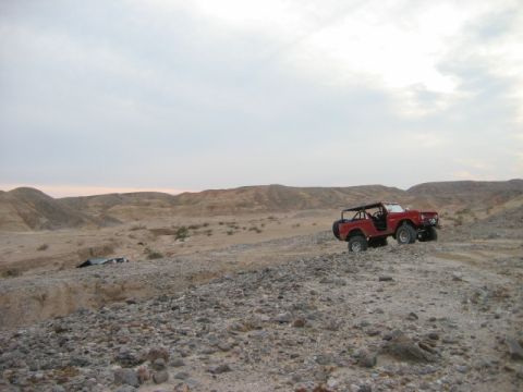 1985 Ford Bronco - A red off-road SUV navigates a rocky desert landscape under a cloudy sky.