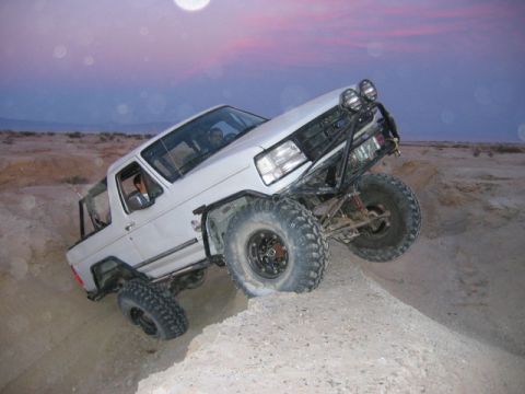 1985 Ford Bronco - White off-road SUV navigates a steep dirt incline under a twilight sky.