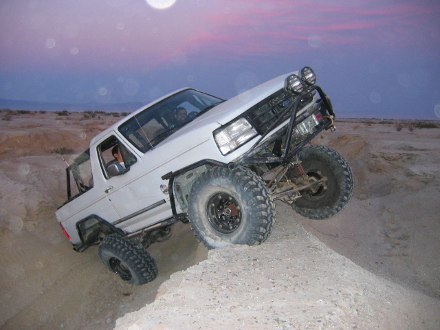 White lifted Ford Bronco on oversized tires climbing a dirt mound at dusk.