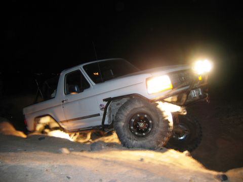 1985 Ford Bronco - White Ford Bronco navigates a dirt incline at night, its headlights illuminating the terrain.