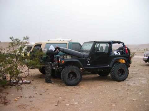 1985 Ford Bronco - Two off-road vehicles are parked in a dusty, outdoor area, with one person attending to the engine of the Jeep.