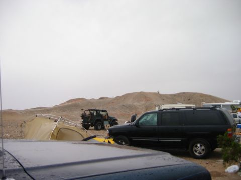 1985 Ford Bronco - A black SUV and a green Jeep are parked in a desert scene with a tent and hills in the background.