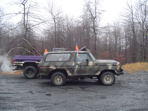 1993 Ford Bronco - Two trucks are parked on a dirt lot with trees in the background. The SUV is camouflaged and the pickup is purple.