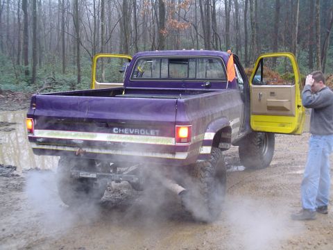 1993 Ford Bronco - A lifted purple Chevrolet pickup truck with bright yellow doors kicks up dust and smoke as it sits in a muddy, wooded area.