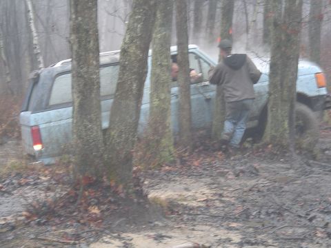 1993 Ford Bronco - A light blue SUV is stuck in the mud in a wooded area, with several people attempting to assist.