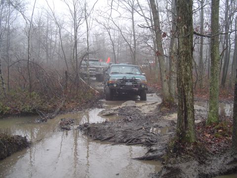1993 Ford Bronco - Multiple SUVs navigate a challenging muddy path in a wooded area on an overcast day.