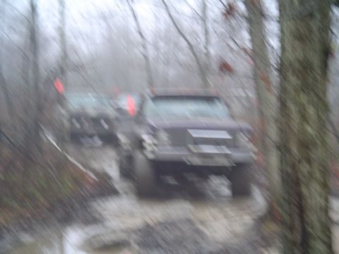 1993 Ford Bronco - Two SUVs navigate a wet and muddy off-road path surrounded by trees.