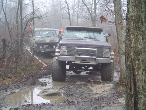 1993 Ford Bronco - Two trucks navigate a challenging muddy path surrounded by trees on a foggy day.