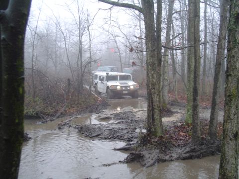 1993 Ford Bronco - Two off-road vehicles navigate a challenging muddy path in a wooded area during overcast weather.