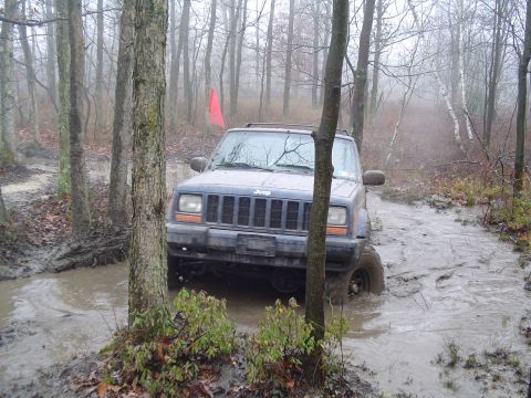 1993 Ford Bronco - A Jeep navigates a waterlogged path in a wooded area on a foggy day.