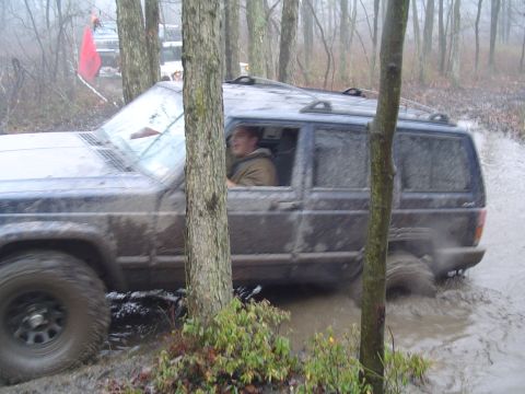 1993 Ford Bronco - A Jeep Cherokee XJ navigates a muddy trail in a wooded area, with another vehicle visible in the background.