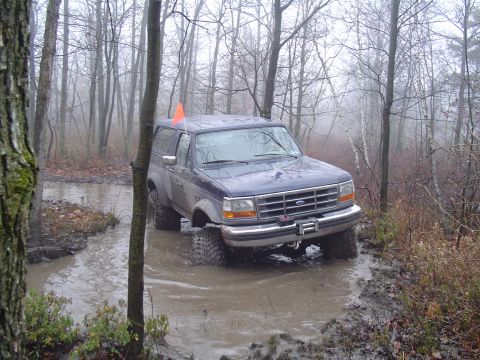 1993 Ford Bronco - A blue Ford Bronco navigates a muddy water obstacle in a wooded area on a foggy day.