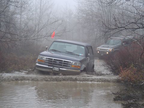 1993 Ford Bronco - Two SUVs navigate a muddy trail on a foggy day, with the lead vehicle partially submerged in water and mud.