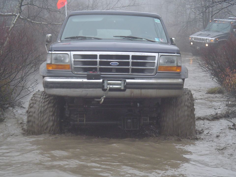 1993 Ford Bronco with large tires driving through a muddy trail, another SUV visible in the background