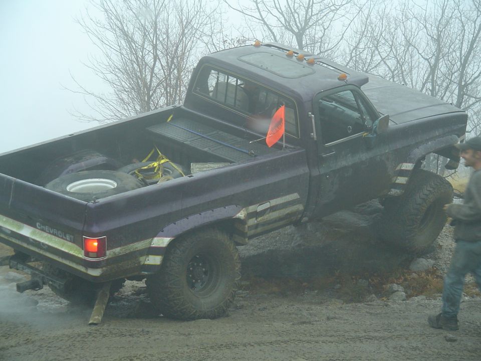Purple pickup truck with lifted suspension stuck in mud, spare tire in bed. Text on rear says CHEVROLET