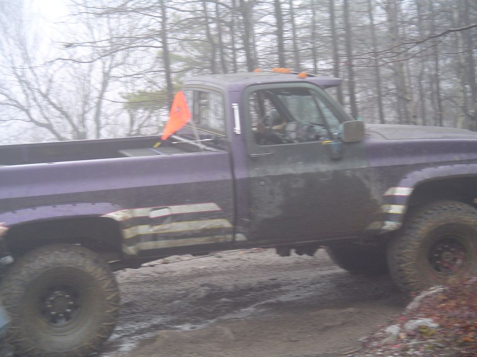 Purple pickup truck with large tires driving on a muddy trail with trees in the background