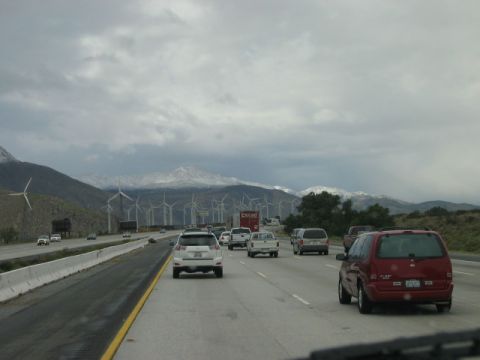 1985 Ford Bronco - A red minivan travels down a highway, with a scenic backdrop of windmills and snow-capped mountains under a cloudy sky.
