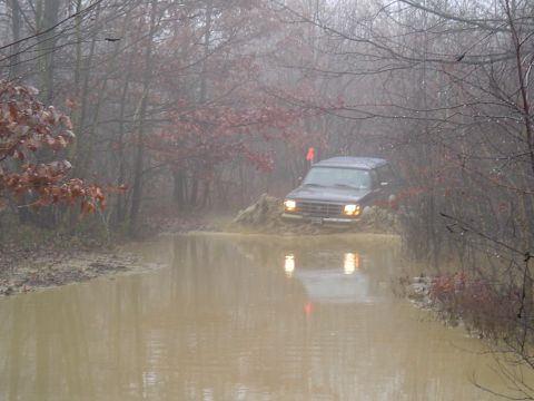 1993 Ford Bronco - A dark SUV navigates a muddy trail, kicking up water and mud.